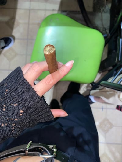 A high-angle shot depicts a hand holding a lit cigar, situated above a green plastic chair, with a portion of a black garment and belt visible in the background. The location is possibly Monte Grande, Argentina. The hand is adorned with a black, open-weave sleeve. The cigar's lit end is held between the thumb and index finger. The background consists of tiled flooring and glimpses of a black shoe. The scene suggests an individual seated on a chair, smoking a cigar in an indoor environment.