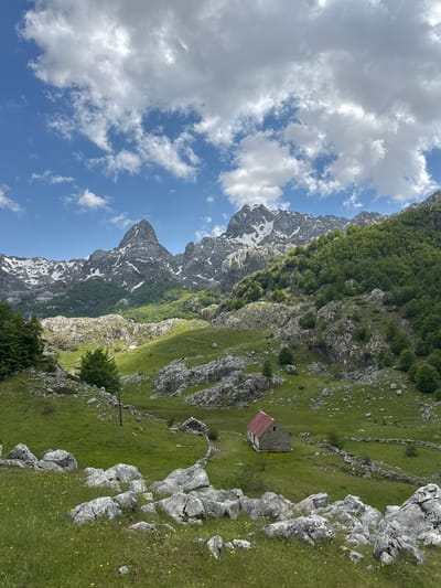 The image depicts a mountainous landscape in Brskut, Montenegro. Rocky, snow-dusted mountains rise on the left under a blue sky with scattered clouds. The right side features a grassy hillside with exposed rocks. A small stone building with a red roof is nestled into the landscape. A low stone wall runs along the hillside. Trees are present among the rocks.