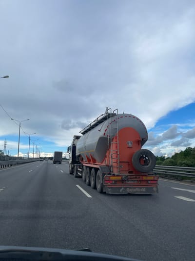 The image shows a grey and orange GuteWolf tanker truck traveling on a highway near Бавыкино, Russia. The truck has a spare tire mounted on the back and ladder access to the top of the tanker. Multiple trucks and cars are driving in the same direction on the highway. Street lights line the side of the road and a forested area is visible on the other side of a barrier. The sky is partly cloudy.
