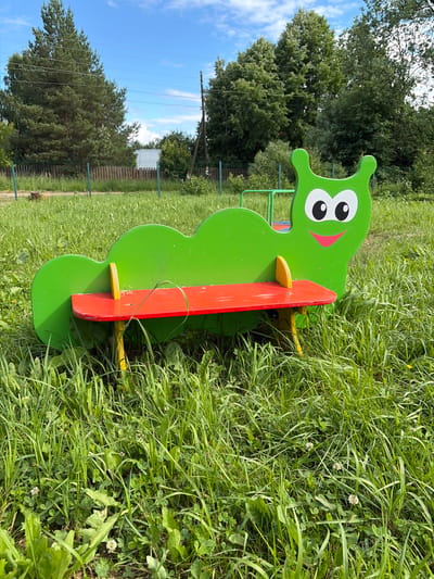 A vibrant, child-friendly bench shaped like a green caterpillar with a red seat is centrally positioned in a grassy area, likely a park or playground in Neklyudovo, Russia. The caterpillar bench features large, cartoonish eyes and a cheerful smile painted on its head. The surrounding environment is characterized by lush green grass, small white wildflowers, and a wooden fence in the background. Trees and a bright blue sky with some clouds complete the outdoor scene. The image appears to have been taken during the daytime on a clear day.