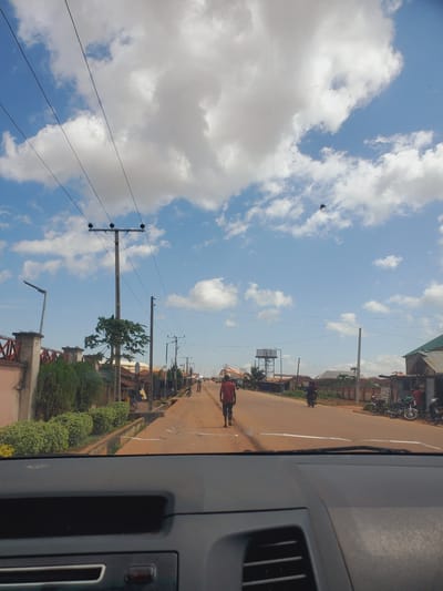 This is a media file taken from inside a vehicle, looking out at a street scene in Bokkos, Nigeria. The image is oriented vertically. The sky is predominantly blue with scattered white cumulus clouds, suggesting a fair weather day. The time of day appears to be daytime, likely mid-morning or afternoon, given the brightness of the sky and the shadows.
The scene depicts a dusty, reddish-brown road lined with buildings and vegetation. Several utility poles with wires stretch across the frame, indicating infrastructure typical of a town or city. On the road, there are a few people and motorcycles. In the foreground on the right, part of the vehicle's interior is visible, including a dashboard element with a vent.
Further down the road, more buildings can be seen, some with corrugated metal roofs. There are also parked motorcycles and some figures walking or riding. Green bushes and trees are interspersed along the roadside. The overall impression is of a busy, everyday street in a Nigerian town. No visible text is present in the image.