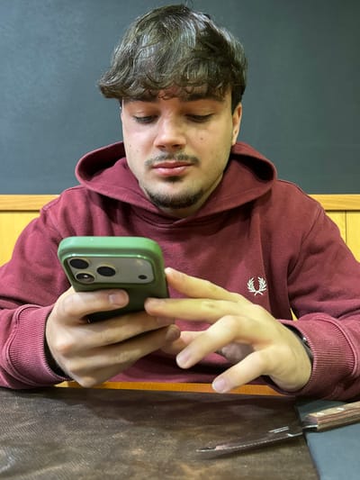 A young male is seated at a table, looking down intently at a smartphone held in both hands. He has short, wavy brown hair, a mustache, and a short beard, and is wearing a maroon hooded sweatshirt with a white Fred Perry logo on the left chest. The smartphone is housed in a textured olive-green case, revealing a triple-lens camera system on its back. His fingers are positioned as if actively interacting with the phone's screen. On the dark brown table surface to his right, a steak knife with a light brown wooden handle and a serrated blade is visible. The background consists of a lower light brown wooden paneling and an upper dark, possibly black, wall. The scene is located within a commercial establishment, likely a restaurant or cafe, in Peso da Régua, Portugal.