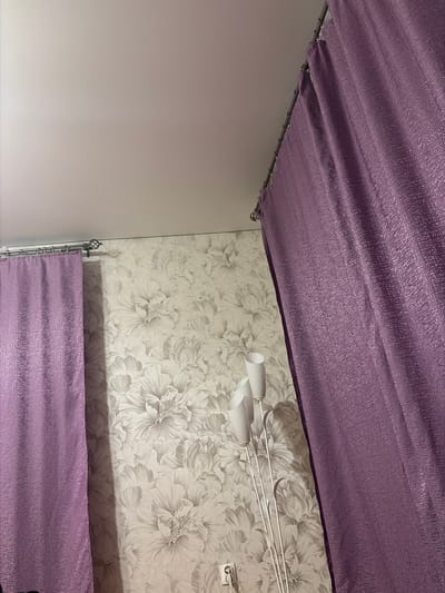The image depicts an interior scene with floral wallpaper and purple curtains. A white floor lamp with multiple light fixtures is partially visible on the right side of the image. The ceiling is white and appears to be a plain surface. The image does not provide any contextual information about its location in the city of Baymak, Russia, or any details regarding people, actions, or interactions.