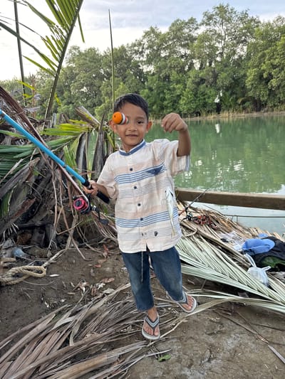 A young boy is standing by a body of water, likely in Grong Grong Capa, Indonesia. He is holding a fishing rod with a bobber on his eye and a small fish in his hand. The boy is wearing a striped shirt, jeans, and flip-flops. The environment is natural, with trees and vegetation surrounding the water. There are also some discarded palm fronds and a blue cap visible on the ground. The lighting suggests it is either early morning or late afternoon.