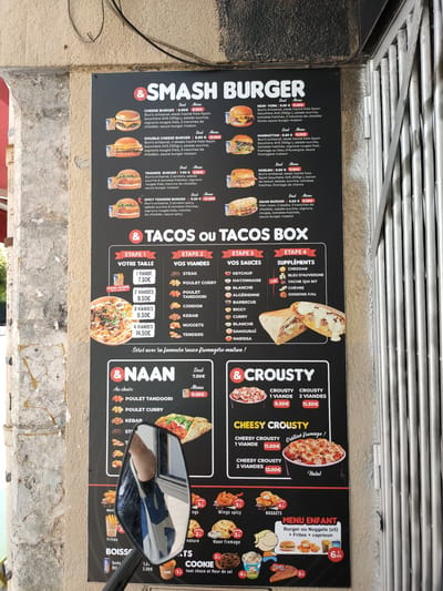 This media file is a photograph taken at an outdoor food stall in Perpignan, France. The main subject is a large, black menu board displaying various burger, taco, and naan options. The menu is divided into sections like "SMASH BURGER," "TACOS OU TACOS BOX," "& NAAN," and "& CROUSTY." Each item is accompanied by a description, price, and often a picture of the food.
The setting appears to be a street-side vendor, with a textured, weathered stone wall on the left and a metal grate structure on the right, suggesting a rustic or urban environment. A motorcycle's side mirror is prominently reflected in the center of the image, showing a person wearing a blue and white t-shirt. This reflection partially obscures the lower section of the menu board, specifically the "NAAN" and "CROUSTY" sections, and some of the food pictures at the bottom.
Visible text includes the names of burger and taco combinations like "CHEESE BURGER," "NEW-YORK," "SPICY TENDERS BURGER," and the process for building a "TACOS BOX" (ETAPE 1-4: VOTRE TAILLE, VOS VIANDES, VOS SAUCES, SUPPLÉMENTS). Prices are clearly listed in Euros (€). Other text indicates "MENU ENFANT" with a price of 6.00€. The overall impression is a vibrant and casual food service operation, likely during daytime given the lighting.