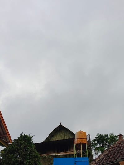 The image displays a static, upward-angled view of a residential or semi-urban scene in Kyauk Hsoke, Myanmar, under a uniformly overcast, light gray sky. In the upper two-thirds of the frame, the sky dominates, showing no distinct clouds or direct sunlight. The lower third features architectural elements and vegetation.
On the left, a partial terracotta-colored tiled roof with an orange-brown underside is visible, extending from the bottom-left corner. Below it and to the right, a dense, dark green tree with textured foliage obscures a portion of the scene.
Centrally located, a building with a steep, dark, possibly corrugated metal gable roof featuring a decorative finial at its apex is prominent. The visible side of this structure below the roof appears to be constructed of dark wood or similar material, possibly with vertical slat or bamboo-like textures. A bright blue wall or structure is partially visible at the base of this building.
To the right of the central building, an orange-yellow, weathered cylindrical water tank is elevated on a light green or metallic support structure. Further to the right, another section of a tiled roof, similar in color and texture to the one on the left, is visible in the lower-right corner. Behind the water tank and extending to the right, parts of a green tree with lighter foliage are present. No people, active subjects, or discernible actions or interactions are present in the scene.