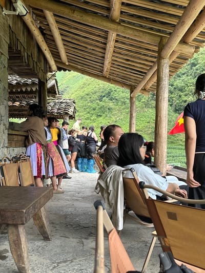 The image shows a group of people, including Vietnamese people in traditional dress, resting on a stone-paved ground under a wooden structure with a bamboo roof, possibly in Yên Minh Commune, Vietnam, given the traditional clothing. The people are seated in chairs or on the ground. A Vietnamese flag is visible in the background of lush green mountains. A security camera is also visible near the structure.