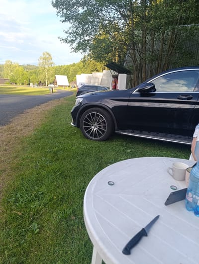 In a grassy outdoor area, possibly a campground or park, a black Mercedes-Benz SUV is parked on the right side of the frame. The vehicle has a sleek design with chrome accents along the bottom. In the foreground, a white plastic table is set up on the grass. On the table, there is a white mug, a dark-colored smartphone, a blue bottle of water, a small knife, and two bottle caps. The background reveals a wooded area with tall trees and a building with a green, corrugated metal exterior. In the distance, there are more trees, rolling hills, and some small structures, including a white triangular-roofed building. The sky is partly cloudy, with a hint of blue. The overall impression is a relaxed outdoor setting.