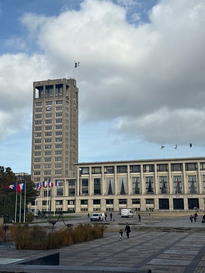 This image captures the Hôtel de Ville (City Hall) complex in Le Havre, France, specifically in the Place de l'Hôtel de Ville, under a partly cloudy sky. The dominant structure is a tall, rectilinear concrete clock tower, approximately 20 stories high, featuring multiple clock faces on its visible sides and numerous evenly spaced, recessed rectangular windows. A French tricolor flag flies from a pole atop the tower.
Adjacent and to the right of the tower is the lower, wider, multi-story main Hôtel de Ville building, constructed in a modernist style with a concrete facade, large recessed window bays, and vertical architectural elements. The inscription "HOTEL DE VILLE" is visible above the ground floor entrance area. On the roof of this building, several flags are displayed on flagpoles, including a blue and yellow Ukrainian flag and other unidentified flags. To the left of the main building, at a lower elevation on the plaza, a series of flagpoles display multiple French tricolor flags.
The foreground and midground consist of a large, paved urban plaza with a geometric pattern of light and dark rectangular tiles. In the immediate foreground, a rectangular planter or water feature contains dry, brown vegetation and several dark, abstract, bird-like metallic sculptures.
Pedestrian activity is observed across the plaza. In the lower right quadrant, a woman wearing dark clothing walks with a small child, who is on a scooter or similar wheeled toy, both moving away from the viewer. Further back, on the plaza near the main building's entrance, two white utility-style vans are parked. Several individuals are gathered around these vehicles, some appearing to be engaged in work or conversation. Additional scattered pedestrians are visible walking across the plaza.