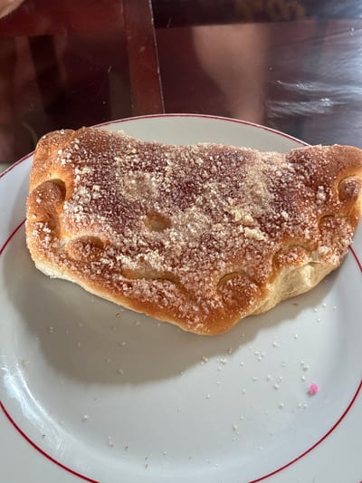 A calzone, topped with coarse sugar and cinnamon, rests on a white plate with a red rim. The calzone has a golden-brown crust and appears to be filled with a dark, jam-like substance. The plate is set on a dark, reflective surface, possibly a table, and a faint reflection of a person is visible in the background. A few stray grains of sugar and a small pink sprinkle are scattered on the plate around the calzone. The scene is lit by overhead lighting, creating subtle shadows. There are no people clearly visible in the image, only a partial reflection. The setting does not provide specific clues to its location in Walasmulla, Sri Lanka.