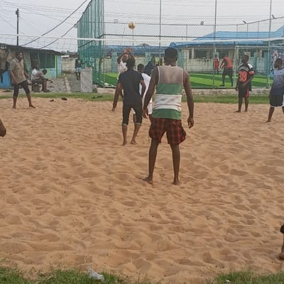An outdoor sandy court in Koko, Nigeria, features a volleyball game in progress under an overcast sky. A yellow volleyball is suspended above a white net. At least ten individuals, predominantly male, are visible. In the foreground, a barefoot man with his back to the camera wears a green, white, and red plaid tank top and red plaid shorts. On the sandy court, multiple barefoot players are positioned; one man in a dark t-shirt and cropped pants is actively reaching for the ball near the net, while others stand ready. To the left, a player in a light brown t-shirt and cropped pants stands, with black sandals discarded nearby. Behind the net, an artificial turf sports field, enclosed by a green chain-link fence, hosts additional individuals, some appearing to play soccer. A light blue building is visible behind the turf. To the far left, beyond the net and turf, makeshift structures with corrugated metal roofs are present, with two individuals seated nearby. Power lines traverse the upper portion of the frame. Small patches of green grass are visible at the bottom, with a piece of white litter.