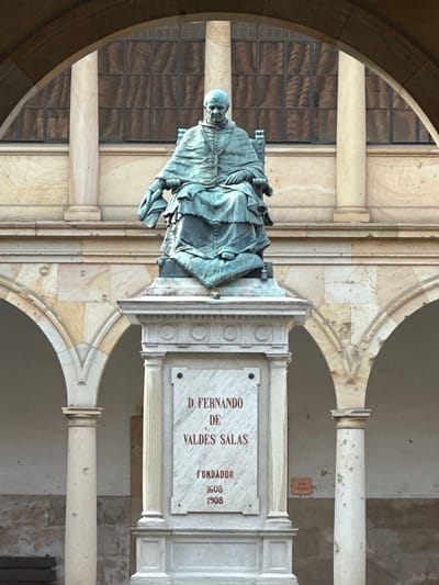 This image shows a statue of D. Fernando de Valdés Salas, the founder of the University of Oviedo in Oviedo, Spain. The statue is positioned on a stone plinth with an inscription indicating his name and the founding date of the university (1608, with a commemorative 1908 date). The statue and plinth are situated within a building, likely a cloister or courtyard of the university, featuring architectural details such as arches and pillars. A window with multiple panes is visible in the background.
