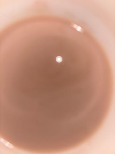 This is a close-up, top-down, abstract shot of a light brown liquid in a container. A bright, circular highlight is visible in the center of the liquid, suggesting a light source directly above or reflected from the surface. The liquid's surface appears smooth and slightly reflective, with subtle variations in color intensity. The outer edges of the image show a lighter, out-of-focus area, possibly the rim of the container. No discernible people, actions, or specific environmental details of Berrechid, Morocco, are visible in this abstract composition.