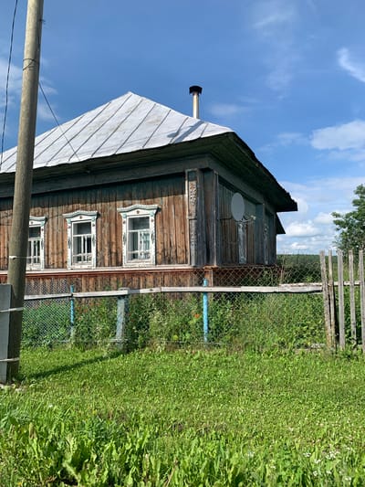 The image shows a weathered wooden house with a tin roof and ornate window trim, set against a blue sky with scattered clouds. A metal satellite dish is attached to the side of the house. In the foreground, a chain-link fence with a wooden section encloses a patch of green grass and overgrown vegetation. A wooden utility pole stands to the left of the house. The scene appears to be in a rural or suburban area.