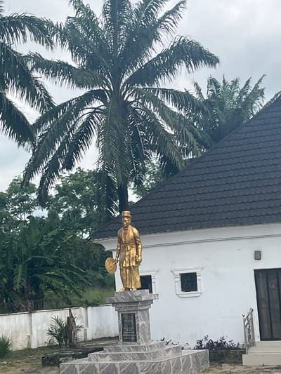 The image, oriented correctly, displays a prominent golden statue positioned centrally. The figure, likely male, is depicted in elaborate traditional attire, including a distinctive hat, and holds a circular object, possibly a ceremonial plate or fan, in their left hand. The statue rests upon a multi-tiered pedestal constructed from grey, patterned stone. A dark plaque is embedded in the front of the pedestal, containing several lines of white text that are not legible due to resolution and distance.

To the left of the statue, a large palm tree with numerous green fronds dominates the foreground, its leaves extending towards an overcast, grey sky. Behind the statue and continuing to the left, dense tropical vegetation, including various green trees and large-leaved plants, forms a lush background.

On the right side of the image, a white building with a dark, possibly tiled or corrugated, roof is visible. The building features two dark-framed windows: a larger rectangular one at the top and a smaller square one below it, accented by a prominent white decorative trim. Below the smaller window, near the building's foundation, are small plants with dark purple foliage. The ground around the monument and building appears to be a mix of concrete or paved surfaces and natural earth.

The scene is a static outdoor shot captured during daytime under an overcast sky, presenting a cultural monument in a tropical environment. The location is Aghalokpe, Nigeria. No specific activity or event is taking place, and no emotions are discernible.