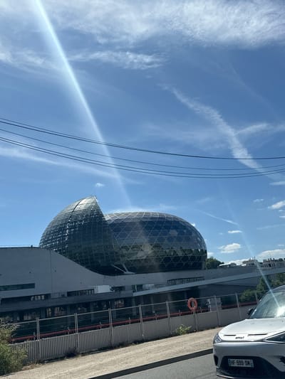 The image shows the Seine Musicale building in Boulogne-Billancourt, France. The architectural complex includes a large glass-covered sphere, which is the main concert hall. A section of a white car is visible in the upper right corner. Utility lines cut diagonally across the top-left portion of the sky, which is otherwise blue with wispy clouds and contrails. A construction barrier runs along the edge of a sidewalk in front of the building.