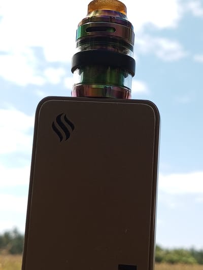 A vape device, featuring a brown box mod with a stylized triple-line logo on its upper left face, is positioned against a backdrop of a partly cloudy sky. The device is topped with a rainbow-colored atomizer tank, secured by a black rubber ring. The background includes blurred vegetation, possibly in a field. The location is identified as Tanjongnie, Indonesia.