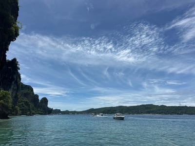 This image depicts a serene coastal scene in Haad Hin Khom, Thailand. The foreground features clear, turquoise water with gentle ripples, and a white rope is visible stretching across the bottom of the frame, suggesting the perspective is from a boat. To the left, a dramatic, lush green cliff face rises from the water, its rocky texture and vegetation indicative of a tropical environment.
In the middle ground, several speedboats are navigating the calm sea. One boat, white with a dark trim, is prominently positioned on the right, while others are further in the distance. The background is dominated by a chain of densely forested hills that form the coastline. The sky above is a vibrant blue, adorned with wispy, white cirrus clouds that create a dynamic pattern. The overall impression is one of a tranquil and picturesque tropical paradise.