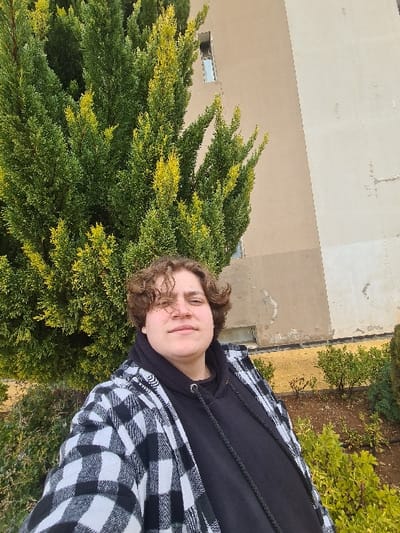 A person stands outside, framed by a building and greenery. The person wears a black hooded sweatshirt under a white and black checkered flannel shirt. They have curly brown hair and a relaxed expression. Lush green bushes and trees with hints of yellow surround them on one side. A tall, light-tan building forms the backdrop on the other side.