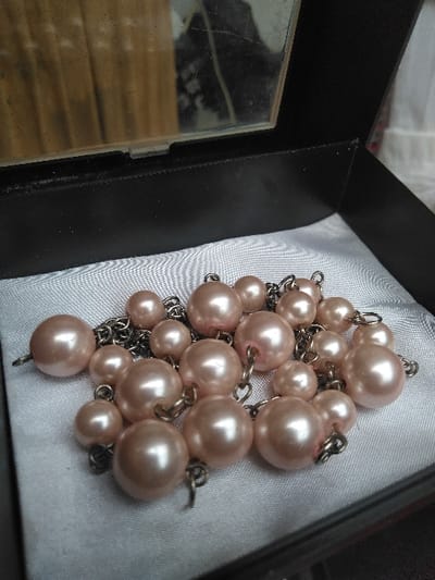 A black, open jewelry box contains approximately 25-30 light pink/peach colored, spherical faux pearl beads of varying sizes, ranging from approximately 0.5 cm to 1.5 cm in diameter. These beads are interconnected by small, dark metal rings and rest on a white, satin-like fabric lining the bottom of the box. The interior of the box lid features a rectangular mirror, reflecting a blurred background consisting of a light brown, vertically textured surface and an indistinct dark shape. The mirror shows signs of minor scuffs or dust. A blurry white background is visible to the upper right of the open box. The scene is located in Лебяжье ауылдық округі, Kazakhstan.