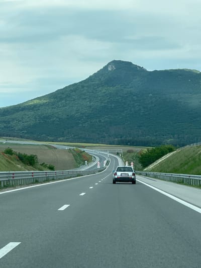 The image shows a silver vehicle driving on a highway near Buhovtsi, Bulgaria. A mountain with dense vegetation dominates the left side of the frame. The highway curves with several warning signs visible. The sky is overcast with a faint blue hue. The road has lane markings and guardrails on both sides.