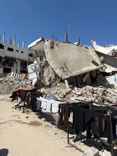 The image depicts a scene of extensive urban devastation in the city of Jabalia, Palestinian Territory, under a clear blue sky. In the foreground, a utilitarian clothesline, fashioned from dark metal bars, spans the lower frame, holding numerous items of recently washed laundry including dark-colored shirts, pants, and lighter-colored shirts and undergarments, hanging to dry. To the left, a folded brown and cream-colored blanket rests on a piece of debris near the ground. The immediate ground consists of compacted dirt and scattered concrete fragments. The midground and background are dominated by the remains of multi-story concrete buildings that have suffered severe structural damage. A prominent, large curved concrete slab, detached and fractured, leans diagonally amidst an extensive pile of rubble in the center-right, exposing internal reinforcing steel bars. To the left, a multi-story building stands partially intact but significantly damaged, with large sections of its facade missing, revealing exposed interior floors, concrete columns, and rebar. No individuals are visible in the scene. The area is illuminated by direct sunlight, casting distinct shadows from the clothesline and debris.