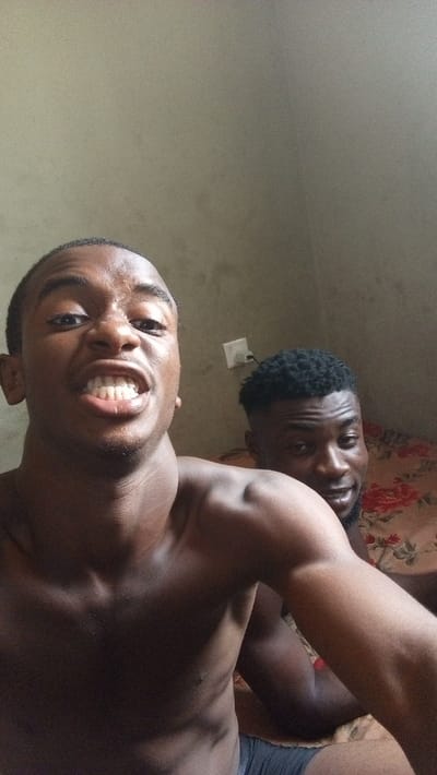 Two dark-skinned men, shirtless, are in a room. The man in the foreground is closer to the camera, baring his teeth. His facial expression is intense. He has a low haircut. Behind him, a second man with a close-cropped afro peeks from behind him with a faint smile. A floral patterned bedspread is visible. An electrical outlet is seen on the wall in the background.