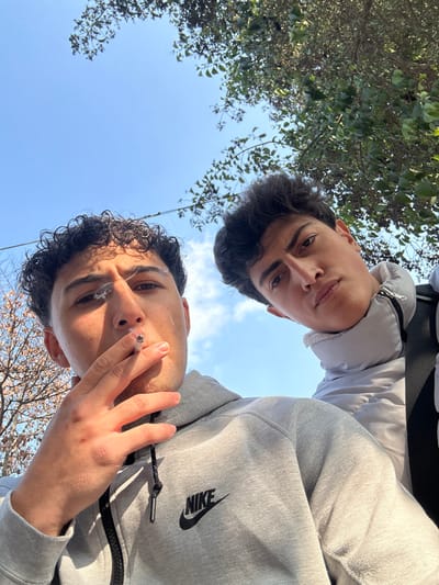 A low-angle outdoor photograph depicts two young males under a clear blue sky with tree branches visible. The scene is located in Al-Qaboun Municipality, Syria.

In the foreground, slightly to the left, a male with dark, curly hair wears a grey full-zip hoodie featuring a black Nike swoosh logo on the left chest. He holds a lit cigarette between the index and middle fingers of his right hand, positioned near his mouth, with smoke actively rising. He looks slightly towards the camera.

Behind and to the right of the first male, a second male with dark, wavy hair is partially visible. He wears a light grey puffer jacket with a visible zipper and a black strap, possibly from a backpack, over his right shoulder. He looks directly at the camera with a serious expression.

The sky is predominantly clear blue, visible in the upper center. Green-leafed tree branches are present in the upper right, while bare or sparsely leafed branches are visible in the upper left. The lighting indicates bright daylight conditions.