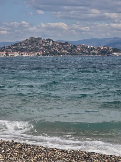 The image shows a view of the town of Astros, Greece, from the sea. In the foreground, there is a pebble beach with gentle waves lapping the shore. The water is a deep blue-green color. In the middle ground, the town of Astros is visible, built on a hillside overlooking the sea. The houses are white with red tile roofs, and there is a castle on the highest point of the hill. In the background, there are mountains. The sky is blue with scattered white clouds.