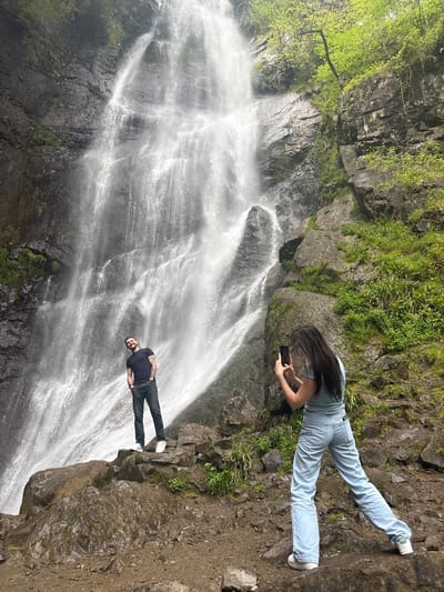 The image captures two individuals admiring a large, powerful waterfall in a natural, lush environment. The scene is presented in landscape orientation, with the waterfall dominating the left and central parts of the frame, cascading over dark, rugged rock faces. The water appears white and misty from its force.
On the right side of the image, closer to the foreground, are two people. A woman with long dark hair, wearing a light blue t-shirt, light blue jeans, and white sneakers, is positioned higher up on a rocky ledge. She is leaning forward, holding a black smartphone with both hands, seemingly capturing a photo or video of the waterfall or the person below. Further down and closer to the waterfall, a man with a beard, dressed in a dark t-shirt, dark jeans, and white sneakers, stands on a rocky outcrop. He is facing the viewer and smiling, with his body slightly turned towards the waterfall.
The surrounding environment consists of steep rocky cliffs and slopes, richly covered with green moss, vibrant green foliage, and small trees, especially visible in the upper right and along the top edges. The ground where the individuals are standing is rocky and earthy. The lighting suggests a bright day. There is no visible text in the image. The setting conveys a sense of natural beauty and adventure, typical of locations like Zeda Makhuntseti, Georgia.