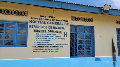 The image displays the exterior wall of a building identified as a hospital in Tchere, Democratic Republic of the Congo. The wall is painted in two distinct horizontal sections: a light beige or cream color on the upper part and a bright blue band at the bottom. Two windows with robust blue frames and security grates are prominent, reflecting the blue sky and white clouds.

A large sign, painted directly onto the upper wall in bold blue lettering, identifies the facility. It reads: "DPS ITURI," "ZONE DE SANTE DE RWAMPARA," and "HOPITAL GENERAL DE REFERENCE DE RWAMPAI." Below this, a list of "SERVICES ORGANISES:" is provided, including "CONSULTATION," "MEDECINE INTERNE," "GYNECO OBSTETRIQUE" (Obstetrics and Gynecology), "CHIRURGIE" (Surgery), "IMAGERIE" (Imaging), and "LABORATOIRE -PEDIATRIE" (Laboratory - Pediatrics). An "H" is painted next to the "SERVICES ORGANISES" heading. The sign also details "HEURES DE SERVICES" (Service Hours): "JOURNEE: 8HOO A 16H59" (Day: 8:00 AM to 4:59 PM) and "NUIT: 17HOO A 08HOO" (Night: 5:00 PM to 8:00 AM).

Above the windows, decorative patterned ventilation blocks are visible. The building features a corrugated metal roof, from which a modern solar-powered floodlight is mounted on the right side, and a simple bare light bulb hangs near the center. A black, pointed object, possibly a furled flag or banner, hangs vertically on the wall to the right of the sign. The scene is brightly lit, indicating daytime, with clear weather. No people are visible, but the detailed sign clearly communicates the building's function as a general referral hospital offering a range of medical services. A watermark "Joseph Kisuki" is present in the lower right.