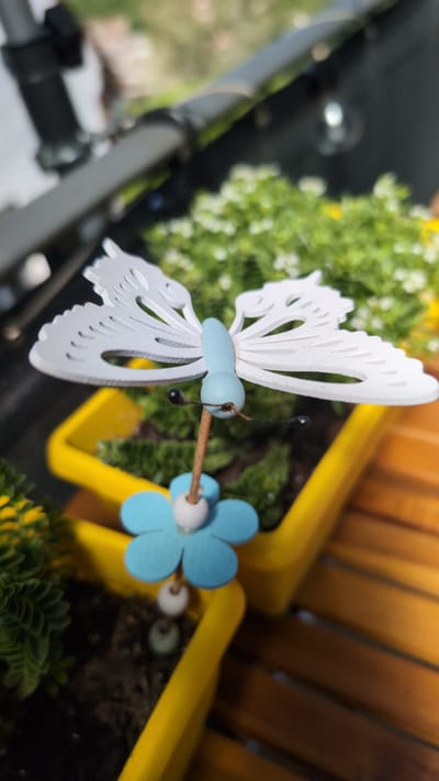 A close-up, low-angle shot focuses on a white decorative butterfly with a blue body and light blue flower accents, mounted on a wooden stick. The butterfly is positioned in front of a yellow planter filled with greenery, possibly moss or small plants. In the background, out of focus, is a blurry gray object, possibly part of a stroller or chair, and a wooden slatted surface. The image appears to be taken outdoors, with natural light. There is no indication of the location being Triberg im Schwarzwald, Germany.