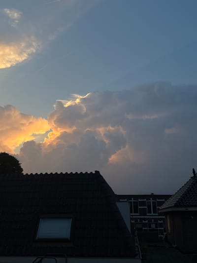 This is a vertically oriented photograph of a sky with dramatic clouds and parts of buildings in Putten, Netherlands. The sky is a deep blue, with a few thin white clouds and contrails visible. Towards the right side of the frame, a large formation of cumulus clouds dominates the sky, catching the warm light of the setting sun, which gives them an orange and yellow glow. The clouds appear dense and textured, suggesting potential for rain or a storm.
In the foreground, parts of two buildings are visible, silhouetted against the bright sky. The building on the right is a dark, tiled roof with a single window. The building on the left has a dark roof and some architectural details suggesting it might be part of a larger structure. The overall impression is one of a dramatic evening sky. The light suggests it is late afternoon or early evening. No people are visible. There is no visible text in the image.