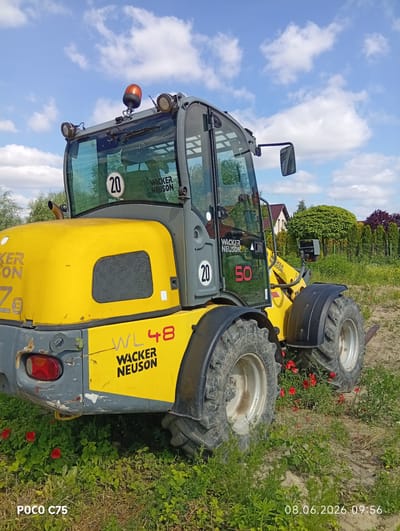 The image shows a yellow Wacker Neuson WL 48 wheel loader parked outdoors on a grassy area with red poppies. The loader is partially obscured by vegetation. The sky is blue with scattered white clouds. In the background, there is a fence and trees, with houses visible beyond them. The timestamp "08.06.2026 09:56" and "POCO C75" are visible in the bottom right and bottom left corners of the image, respectively. The location is identified as Kiełczów, Poland. There are no people visible in the image. The loader appears to be in good condition, with some minor scuff marks on the gray parts of its body.