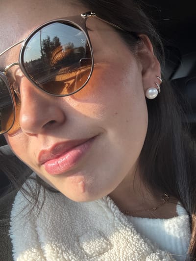 A close-up of a woman's face, angled sideways, fills the frame. She wears round sunglasses reflecting a street scene. Her skin is fair and illuminated by sunlight. A large pearl earring adorns her ear, with smaller rings above it. Dark hair frames her face. She wears a textured white top. A delicate gold necklace with small charms is visible near her collarbone.