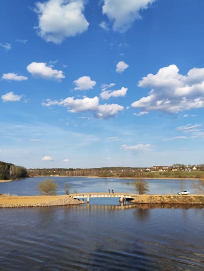 The image depicts a view of a lake with a narrow land passage connecting two sides. A concrete bridge spans a narrow channel in the land passage. Two people are standing on the bridge. A white car is parked on the land passage to the right of the bridge. The lake is surrounded by trees and buildings in the background. The sky is blue with puffy white clouds. The location is Podosinki, Russia.