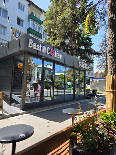The image shows the exterior of a "Best Coffee" shop in the city of Edineț, Moldova. The shop is a modern, glass-fronted structure with white lettering spelling out the business name, accompanied by a graphic of a hand holding a coffee cup and a pink donut. String lights with exposed bulbs are draped across the front of the building and over the adjacent trees. In the foreground, two black circular tables with accompanying chairs are visible on a paved walkway. Beyond the tables, a wooden planter box filled with various green and reddish plants is situated.
The shop's facade features large windows that reflect the surrounding environment, including trees, buildings, and street activity. Through the windows, the interior of the coffee shop can be partially seen, with neon signs and possibly displays of products. On the right side of the building, there is a sign indicating "SCHIMB VALUTAR" with currency symbols for dollars and euros, suggesting currency exchange services are offered. Further down the street, a white car is parked and a green bus is visible, indicating a public street environment. The overall scene is well-lit by daylight, with shadows cast by the trees and the coffee shop structure.