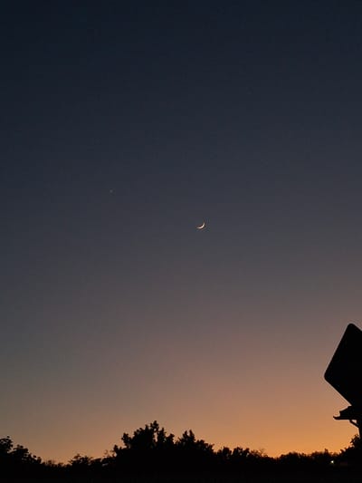 The image depicts a crescent moon visible in the evening sky above Dragomirovo, Bulgaria. The sky transitions from a darker blue-gray in the left portion of the frame to an orange-tinged glow on the right, likely indicative of sunset. Silhouetted trees and an indiscernible object occupy the right edge and upper-right corner of the image, respectively. A dim point of light, possibly a star or planet, is visible beneath the moon.