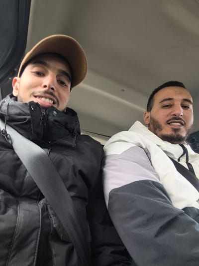 This image captures two young adult males seated inside what appears to be a vehicle's cabin. The specific location within Sabaa Aiyoun, Morocco, cannot be determined from the provided visual information.

The subject on the left is a male with light skin, dark hair, a short mustache, and a beard. He wears a light brown baseball cap, a black puffer jacket, and a grey seatbelt crosses his chest diagonally from left to right. He is smiling, displaying his teeth, and looking directly at the camera.

The subject on the right is a male with light skin, dark hair, and a short beard. He wears a multi-colored puffer jacket with white, light grey, and dark grey sections. He is also smiling, displaying his teeth, and looking directly at the camera.

Both individuals are positioned close to each other, indicating they are likely seated side-by-side. The background is a plain, light-colored ceiling of an interior space. The image is a close-up, seemingly a selfie, with both subjects actively posing for the photograph.