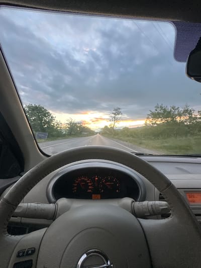 The image shows the view from inside a car driving on a road, likely in the Padarevo, Bulgaria area based on the landscape visible through the windshield. The car's steering wheel and dashboard are visible in the foreground. The dashboard displays a speed of approximately 10 km/h, and the engine is running at around 1000 RPM. The exterior environment features a road stretching into the distance, flanked by trees and fields illuminated by the setting sun or rising sun. The sky is cloudy with varying shades of grey and hints of orange and yellow light.