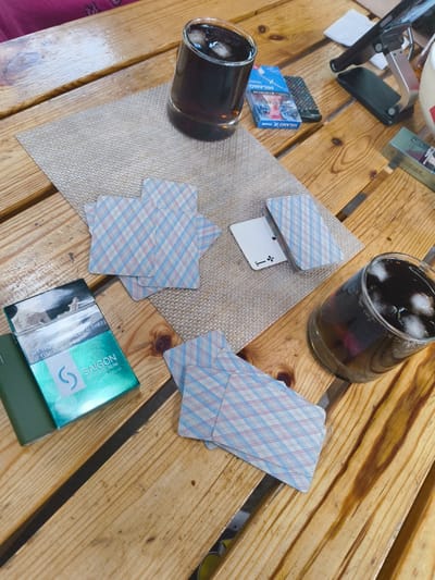 In the city of Востец Снт, Russia, a wooden table is set with items for a casual gathering. Two glasses of dark beverage with ice are present, one in the upper right and another in the lower right. Scattered across the table are several decks of playing cards, some face down and some partially visible. A pack of "SAIGON" cigarettes, colored green and white with Vietnamese text, is open on the left side of the table, with a cigarette pack of "MILANO CHANGE" and a small remote control to its right. A white slip of paper with the number "1" is visible among the cards. A tablet stand is positioned in the upper right of the frame, with a dark-colored tablet device resting on it. The scene suggests a moment of pause during a game or conversation.