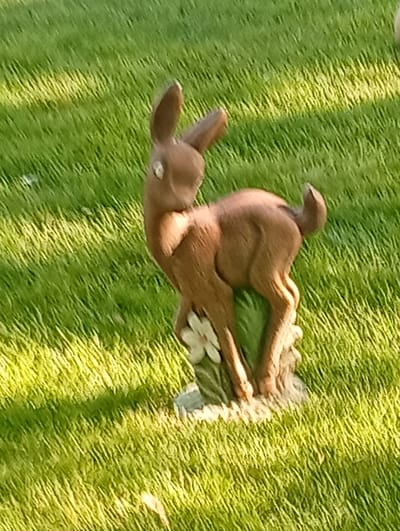 A brown ceramic fawn figurine stands on a green grassy lawn. The figurine is positioned in the center of the frame, facing left and slightly upward. It has large ears, round white eyes, and a short, curved tail. A small white flower is visible at the base of the figurine. The grass is illuminated by sunlight, creating a pattern of light and shadow. The background is entirely composed of this grassy lawn, with no other objects or structures visible. The scene is likely outdoors, in a garden or park setting.