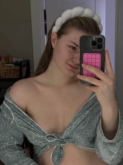 A fair-skinned person with light brown hair is taking a selfie in a bathroom. She is wearing a white, puffy headband and a blue-gray patterned robe tied in front, revealing cleavage. She is holding a gray iPhone with a pink pop-it case. The background is a typical bathroom setting, including a cabinet and a window or mirror reflection. There's nothing to suggest that this picture was taken in Yanga-Yul, Russia.