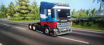 A digital rendering of a Scania truck with a Russian flag-themed paint job is parked on the side of a road in Mokhsogollokh, Russia. The truck is dark blue, white, and red, with wavy lines. It has a chrome grille and bumper, and auxiliary lights on the roof. The truck is viewed from the front-left side, angled towards the viewer. The road is paved and has white lane markings. The surrounding environment is a rural landscape with green trees and grass, under a clear blue sky. A road sign with a yellow triangle is visible on the left. The scene appears to be a still from a video game.