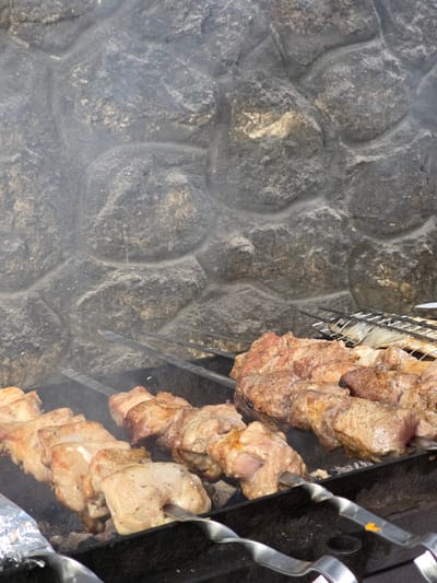 In Pidhorodne, Ukraine, an outdoor barbecue is underway, with several skewers of meat cooking over hot coals. The meat, appearing to be pork or chicken, is arranged in chunks on long metal skewers. The background features a textured stone wall, likely part of an outdoor cooking area or structure. Smoke rises from the grill, indicating the cooking process is in progress. A metal grill rack is visible in the upper right portion of the image, suggesting other items might also be cooking. The overall scene depicts a common method of food preparation in an outdoor setting.