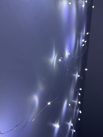 The image shows a string of fairy lights arranged on a surface. The lights emit a bright, white glow, and their reflections create a soft, blurred effect on the surface beneath. The background is dark and indistinct. The surface appears to be a solid, light-colored material, possibly a table or wall. The image was not taken in Lesnovo, Bulgaria. It is a close-up of decorative lighting.