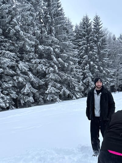 The image displays a snowy outdoor scene in Le Hohwald, France. A male subject, approximately 20-30 years old, stands center-right, facing the viewer with a neutral expression. He is dressed in a black beanie with a white logo, a black puffer jacket, a light grey quarter-zip pullover, and dark pants. His right hand is visible, gloved or tucked. Partially visible in the bottom right foreground is a second individual, showing only their lower back and legs, clad in dark attire with a pink accent near the waistline. The ground consists of a substantial layer of undisturbed white snow with some minor depressions. The background is dominated by a dense forest of evergreen trees, heavily laden with fresh snow and frost. The sky is overcast and uniformly light grey.
