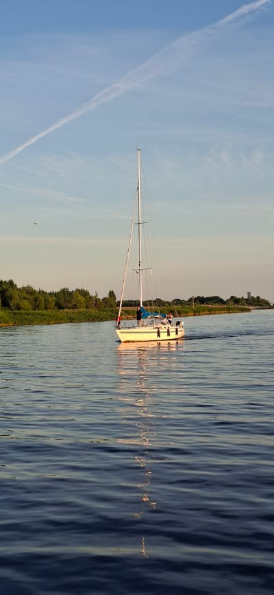 A white sailboat with a tall mast and two people on deck is sailing on calm water near a lush green shoreline under a clear blue sky with a faint contrail. The boat is moving from left to right in the frame, creating a slight wake behind it. The shoreline is lined with dense trees and reeds. The water reflects the sky and the boat, creating a shimmering effect. The scene appears to be set in a natural environment, likely a river or a bay, and based on the context provided, it is in Pärnu linn, Estonia.