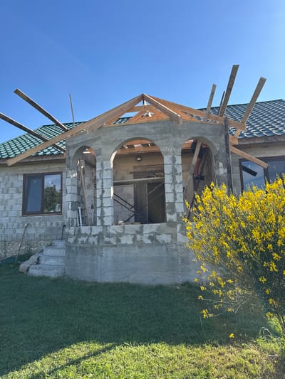 The image depicts a house under construction in Pîrlița, Moldova. The scene is outdoors under a clear blue sky, suggesting fair weather and likely daytime due to the bright sunlight casting shadows and illuminating the grass.
The primary subject is the house, which is in an unfinished state. The exterior walls are constructed from light-colored cinder blocks, and some areas are covered in wet concrete, indicating ongoing building work. A prominent feature is a large, arched opening leading into the house, with wooden scaffolding and framing around it. Another smaller, rectangular window is visible on the lower level of the house. The roof is partially covered with dark, textured tiles, and more wooden beams are positioned around the roofline, suggesting a continuation of the construction process.
To the right of the house, a vibrant bush with numerous small yellow flowers is in bloom, adding a touch of color to the scene. Beyond the bush, a well-maintained patch of green grass slopes upwards. To the left of the house, the sky is visible, a stark contrast to the earthy tones of the construction materials.
The overall impression is one of progress and development. There are no people visible in the image, and no text is present. The setting is clearly a residential area, likely a suburban or rural location, given the presence of both the construction site and the natural elements like the grass and flowering bush.