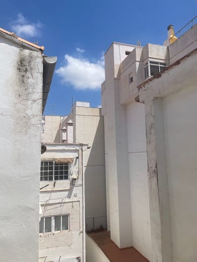 This image captures an outdoor scene with a clear blue sky and scattered white clouds, suggesting fair weather during the daytime. The perspective is looking upwards at the sides of several multi-story buildings. The architecture appears to be residential or commercial, characterized by white and beige facades, multiple windows, and concrete structures.
Several notable architectural elements are visible. On the left, a bright yellow vent protrudes from a building. There are various windows, some with dark metal grids, others with white frames. Antennas are present on some of the rooftops. One building shows signs of wear and tear, with visible cracks and peeling paint on a concrete beam. On the right side of the image, a red-brown colored wall is visible within the structure of one building.
In the foreground, a white-tiled wall with visible cracks and staining runs horizontally across the bottom of the frame. On the far left, the top of a dark grey, curved pipe or chimney is seen on a tiled roof. The overall impression is an urban or semi-urban environment, likely in a Mediterranean climate given the presence of bright sunlight and blue sky. There are no people or active events depicted, focusing solely on the architectural features and the sky. No visible text is present in the image.